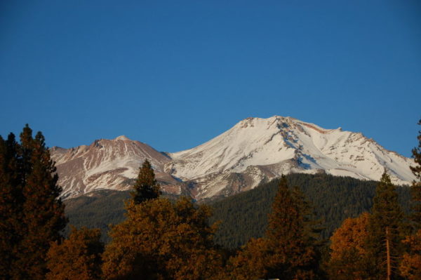 This is a photo of a a view of Mount Shasta in the Fall that has a dusting of snow on the mountain with red and orange leaves on the trees in the foreground.