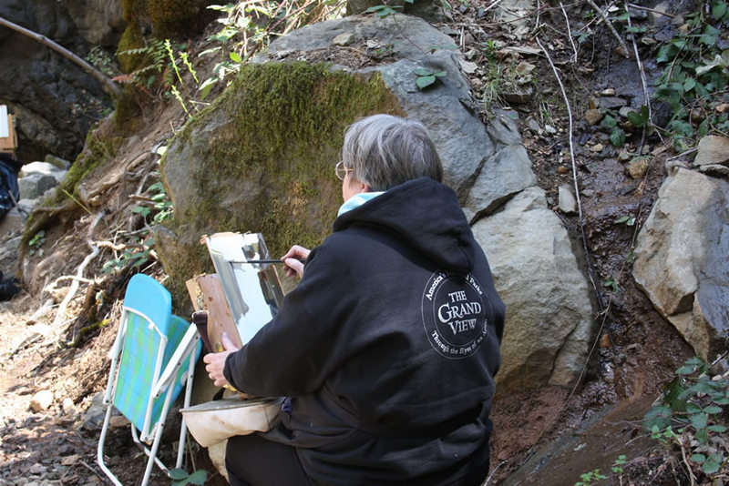This is a photo of Kris Baxter sitting on a rock and painting plein air at Hedgecreek Falls during a Spring painting Workshop.